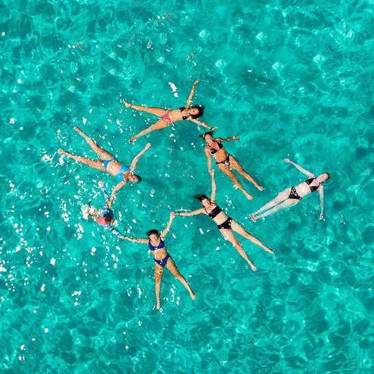 Aerial view of seven people floating on their backs in crystal-clear, bright turquoise tropical water. They are arranged in a loose circle, arms outstretched with some holding hands, forming a starburst pattern. Sunlight glints across the surface of the shimmering water.
