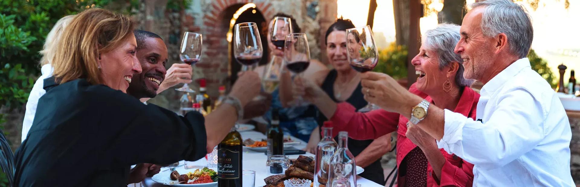 A group of diverse friends laughing and raising wine glasses in a toast around an outdoor dinner table setting.