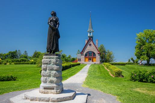 A bronze statue of an Acadian woman in a long dress stands on a stone pedestal in the foreground, with the red and white stone Acadian Memorial Church featuring a tall steeple visible in the background under a clear blue sky. The scene is set in well-manicured grounds with green lawns, trimmed hedges, and a paved walkway leading to the church entrance in Pobomcoup, Nova Scotia.