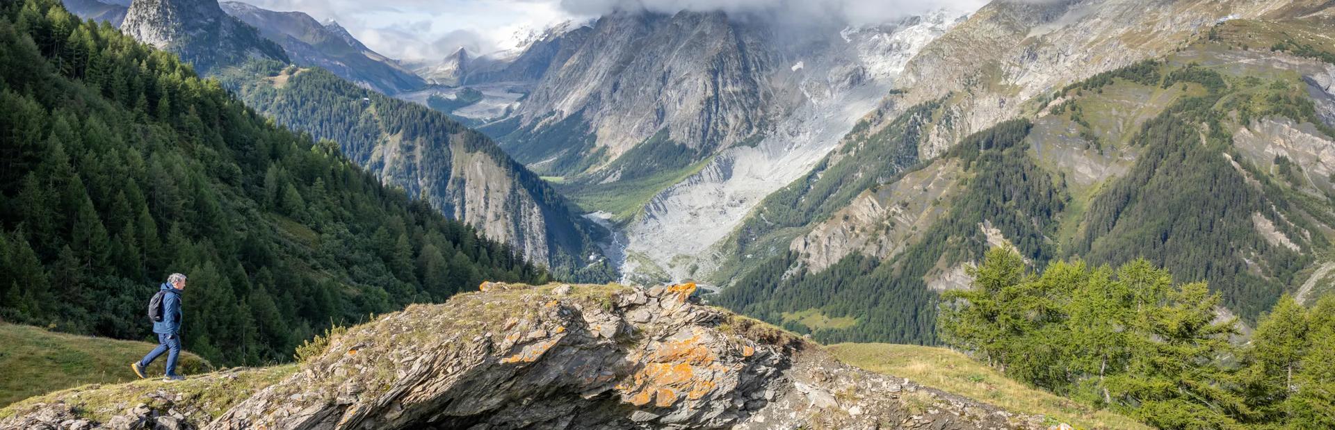 A side view of a hiker with a backpack walking along a grassy mountain ridge overlooking a vast, dramatic valley with steep rocky mountains and forested slopes under a partly cloudy sky.