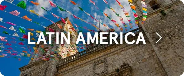 Colorful banner flags strung across a blue sky above a historic stone building and church tower, overlaid with the text "LATIN AMERICA" in bold white letters in the center.